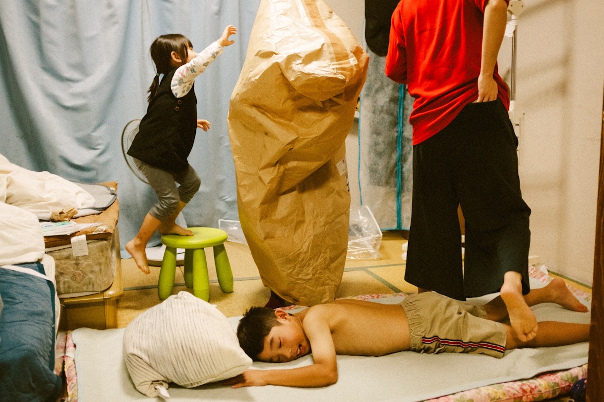 Jun Aihara FUJIFILM X100F撮影の布団にうつ伏せになる上裸の子供とそれをまたぐ大人とその後ろで椅子に登る子供